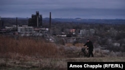 Un bărbat își plimbă bicicleta de-a lungul unei cărări de pe un deal deasupra New York-ului. Luminile din fundal sunt ale Horlivka, un oraș controlat de separatiștii susținuți de Kremlin. Fabrica de fenoli din New York este în stânga.